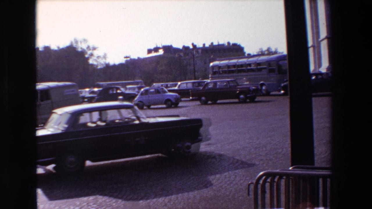 1969 Paris street traffic seen from indoors through a window, with classic cars, a bus, cobblestone paving, and distant buildings or trees.