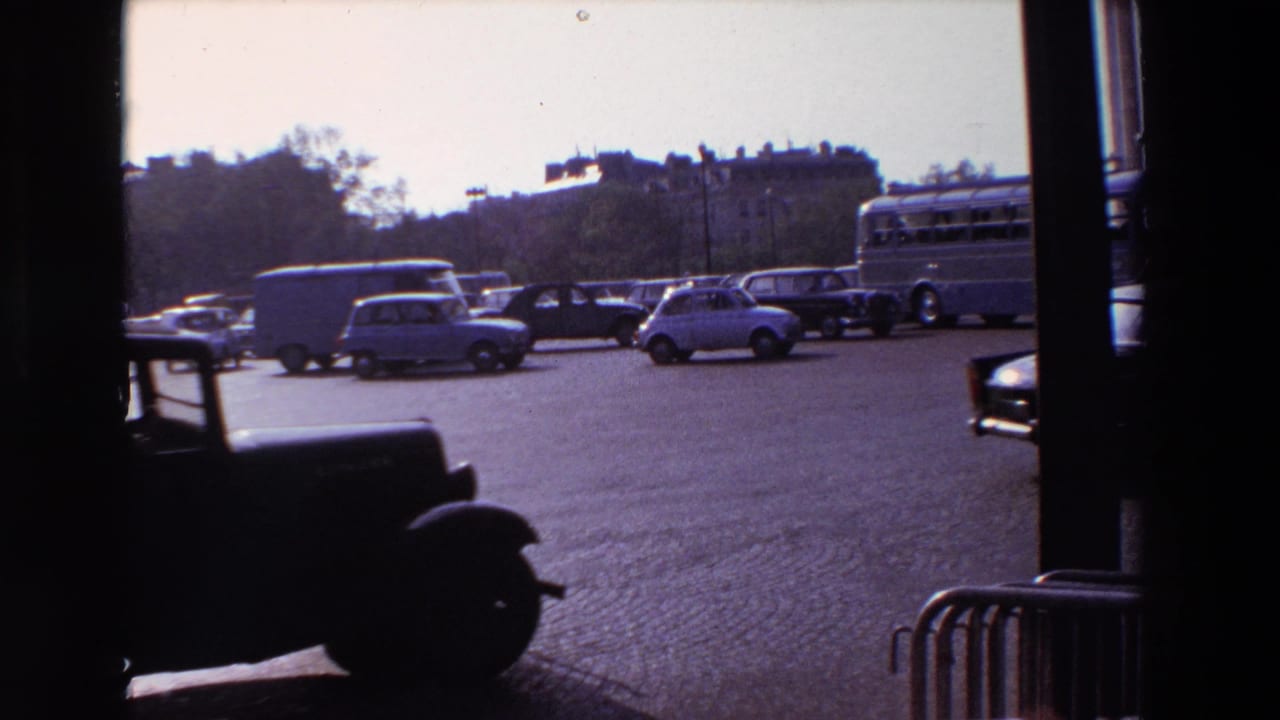 A dark vehicle passes in front of cars and a bus on a cobblestone street.
