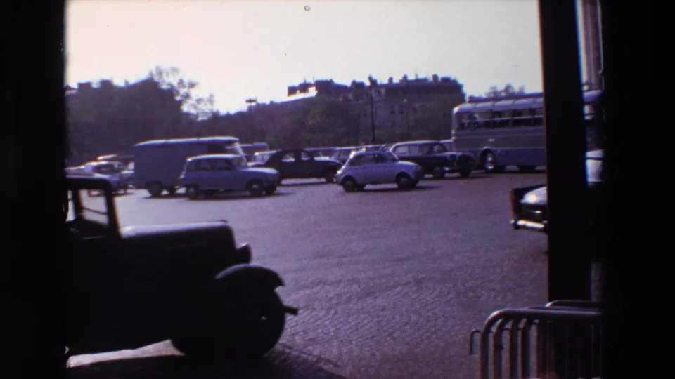 A 1969 Paris street view through a window shows a dark vehicle crossing the foreground, with compact cars, a bus, and cobblestones behind it.