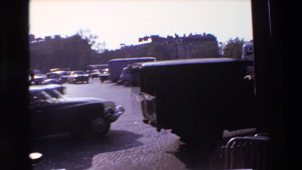 Shadowed 1969 Paris window view with moving cars, a dark van or truck, cobblestone paving, and a line of parked vehicles in the background.
