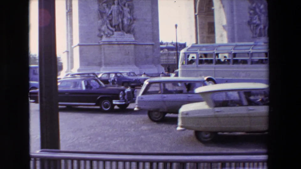 1969 Paris traffic near a large stone monument base, with classic cars, a bus, cobblestones, window framing, and small pedestrians visible at a distance.