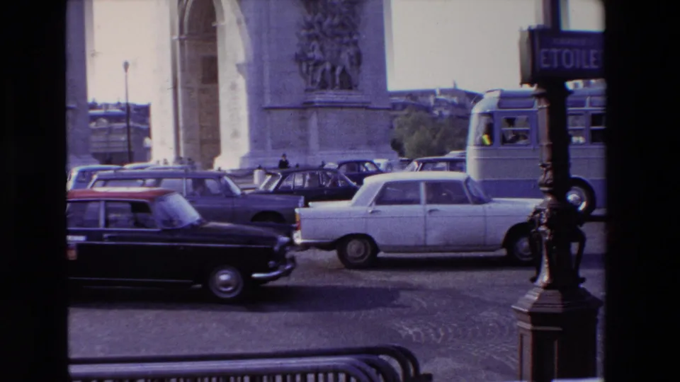 1969 Paris home-movie frame showing a white sedan, darker cars, a bus, and a lamppost near the Arc de Triomphe traffic circle.