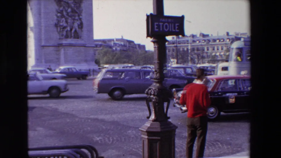 1969 Paris frame showing a pedestrian in a red sweater near a Place de l'Etoile sign, with cars, buildings, and the Arc de Triomphe area behind him.