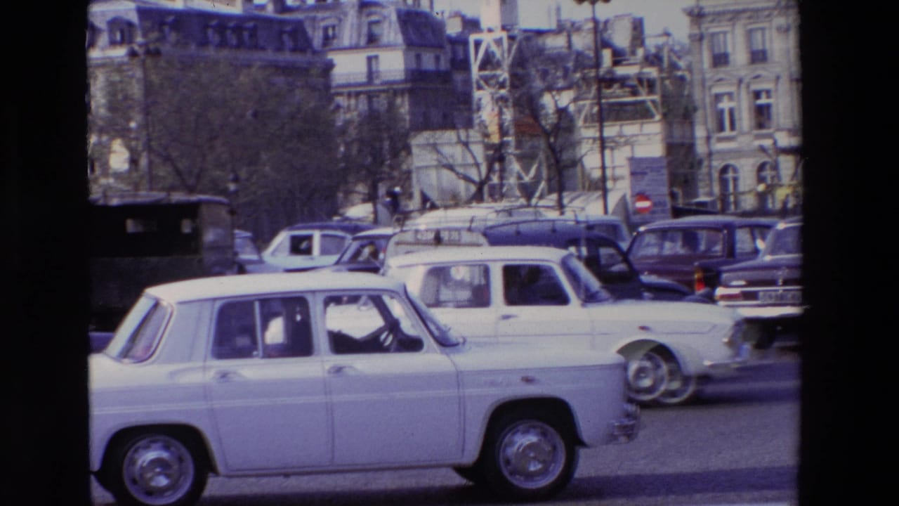 A white Renault 8-style sedan moves across the foreground of a 1969 Paris street scene, with other period cars, bare trees, and mansard-roof buildings in the background.