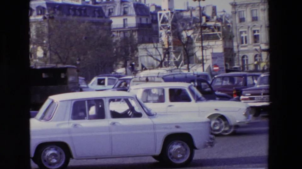 A white Renault 8-style sedan moves across the foreground of a 1969 Paris street scene, with other period cars, bare trees, and mansard-roof buildings in the background.