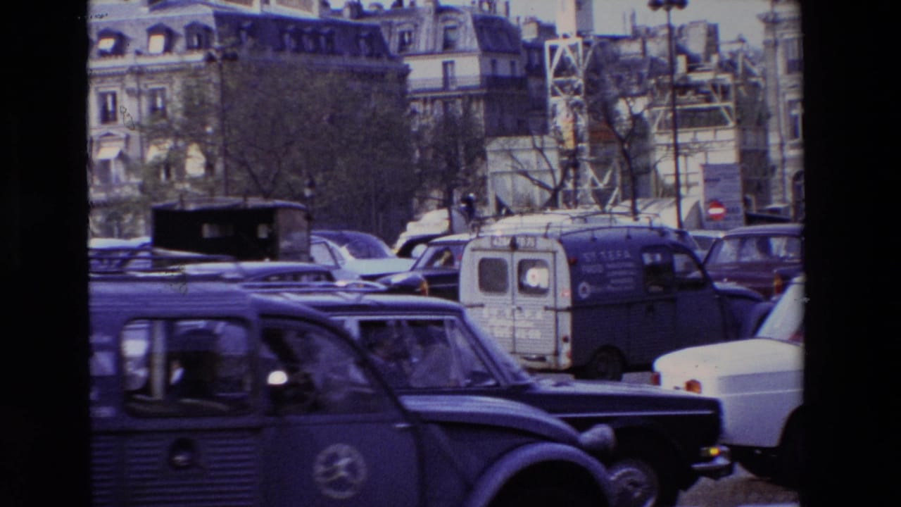 Blue Citroen-style van and dark car moving through dense city traffic near Parisian buildings.