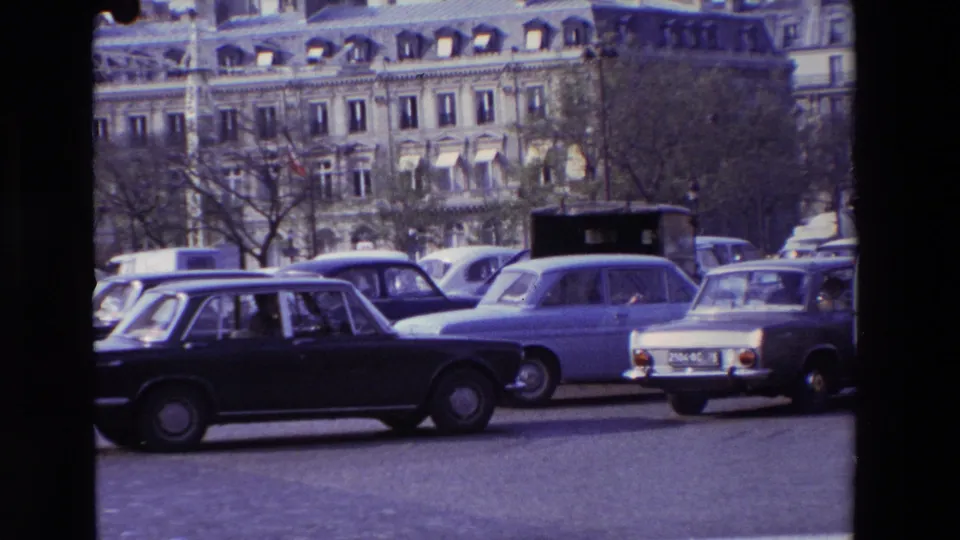 Several 1960s sedans move through a Paris street in 1969, with curbside traffic, bare trees, and Haussmann-style apartment facades filling the background.