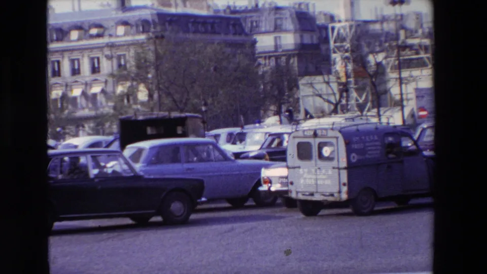 A blue Citroen 2CV-style van and multiple sedans continue through a busy 1969 Paris street scene, framed by bare trees, stone buildings, and a metal structure in the background.