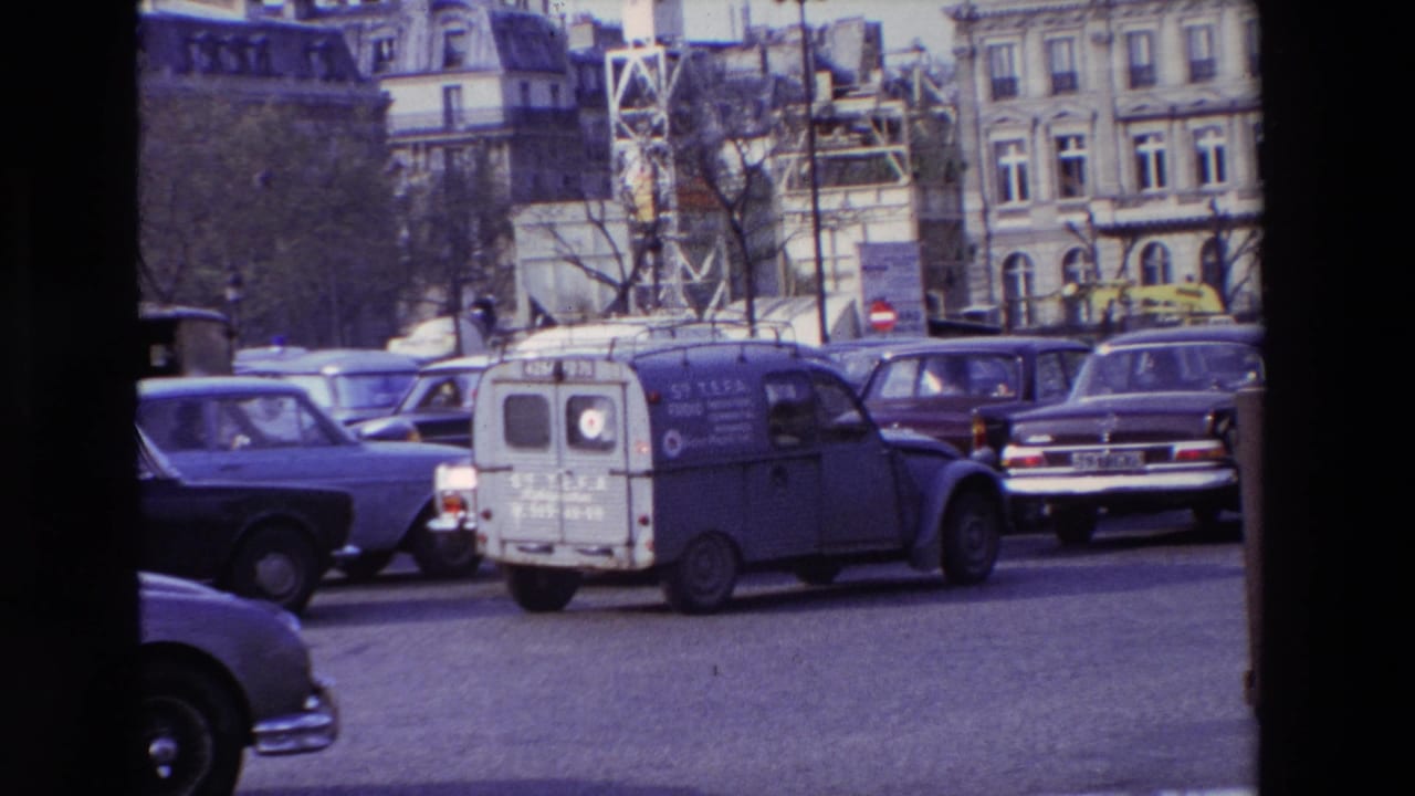 1969 Paris street traffic with a blue Citroen-style utility van among older cars in front of ornate stone buildings.