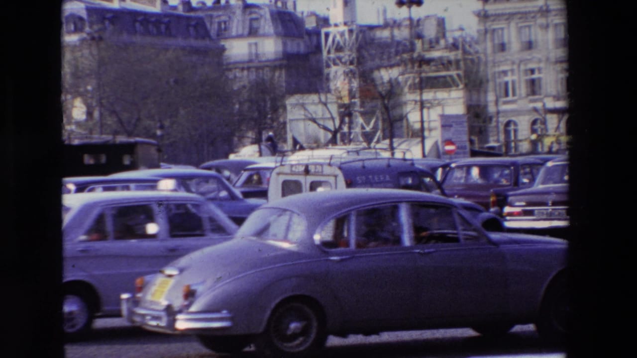 1969 Paris traffic scene with a large dark sedan and other period cars moving past Haussmannian-style buildings.
