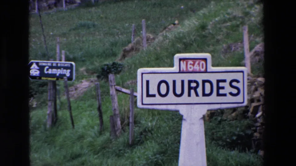 French roadside sign for Lourdes on the N640, set against green hillside, wooden fencing, and a nearby camping direction sign.