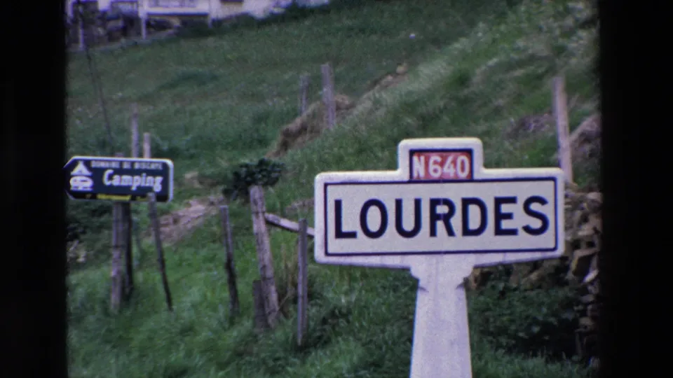 French countryside view with the Lourdes N640 sign prominent in the foreground and a camping direction sign near a grassy fenced slope.