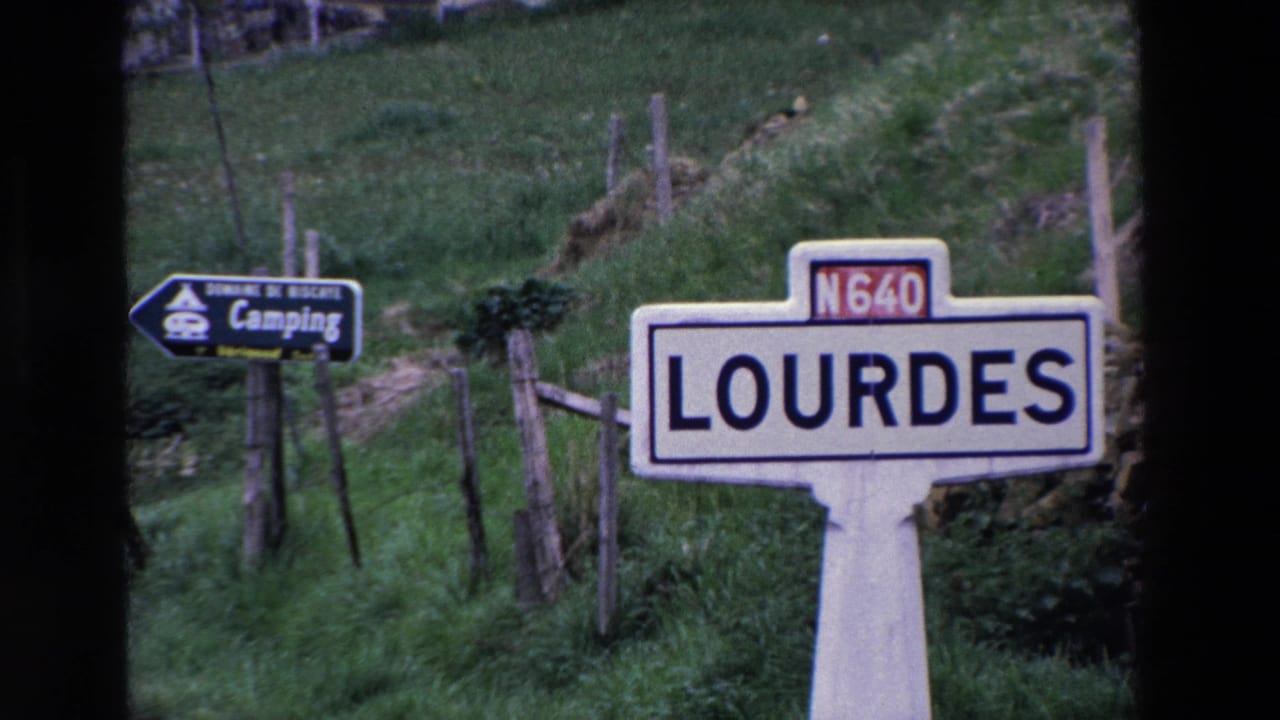 1969 8mm frame of a white Lourdes road sign on the N640 route, with a camping sign, fence posts, grass, and stonework in the background.