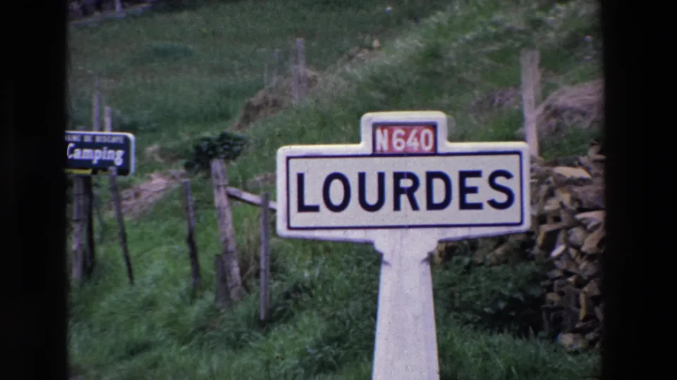 A handheld 8mm view centers the Lourdes place-name sign and N640 marker, with a small camping direction sign and rural hillside behind it.