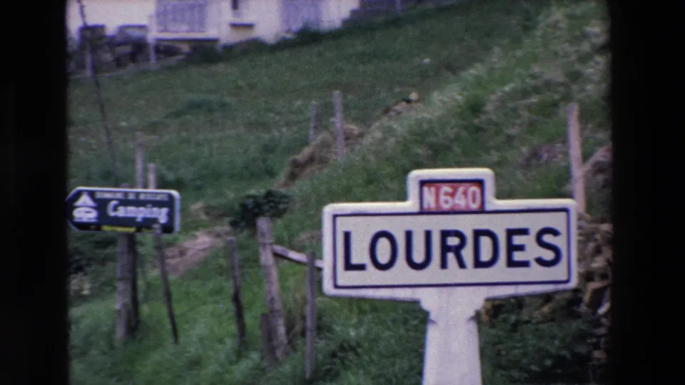 The Lourdes roadside sign remains in view, cropped lower in the frame, with the camping sign, fence line, grass, and hillside visible behind it.