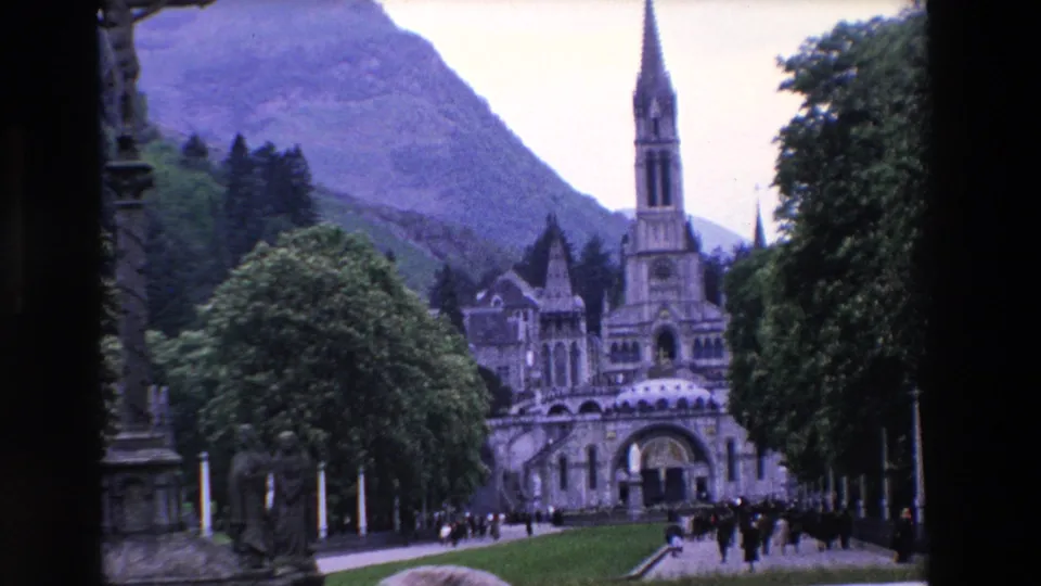 1969 8mm travel frame of the Basilica of Our Lady of Lourdes seen from a distance, with a tall spire, tree-lined approach, mountains, lawn, and visitors on the pathways.