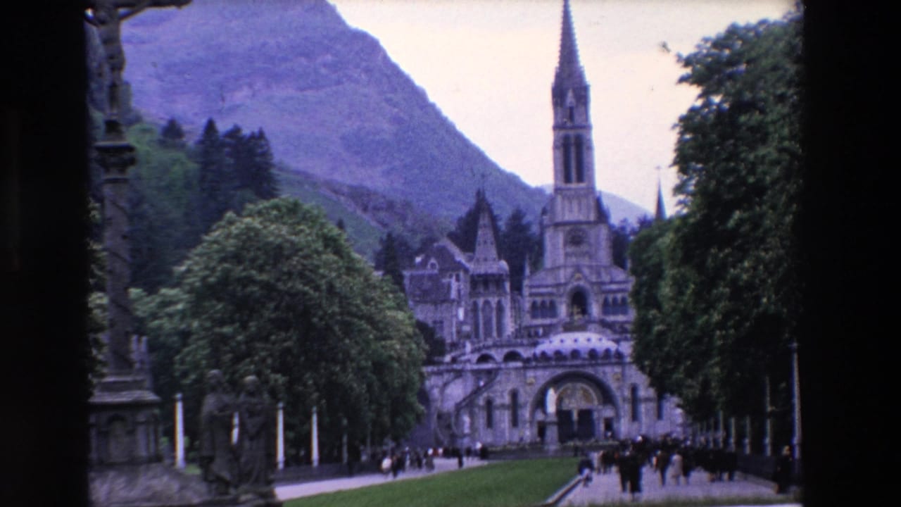 1969 8mm travel footage showing visitors approaching an ornate religious complex in France, with trees, a stone monument, lawns, and mountains in the background.