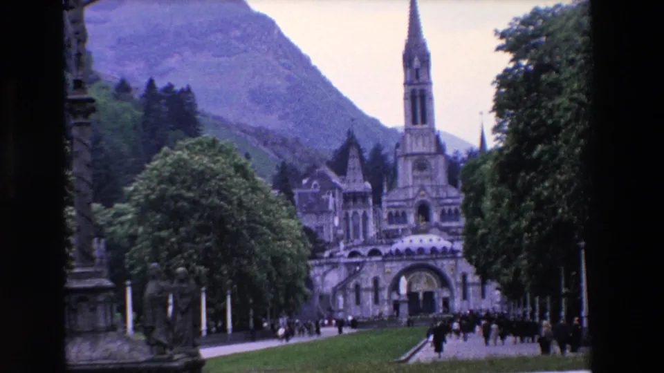 A handheld 8mm frame shows the ornate religious complex from the approach path, with distant visitors, dark trees, green lawn, and a mountain slope behind the spire.
