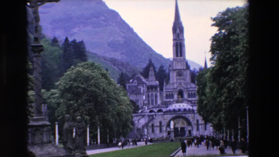 The final still shows visitors dispersed across the approach to the ornate religious complex, with the tall spire, arched lower facade, trees, lawns, and mountains visible.