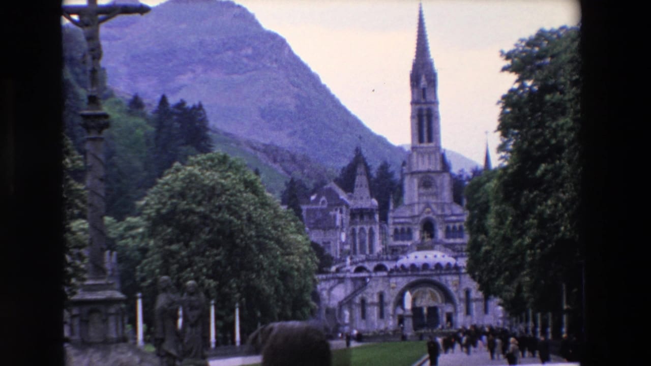 1969 8mm travel footage frame of a French basilica approach, with a tall central spire, stone facade, trees, mountain backdrop, and visitors walking near the entrance.