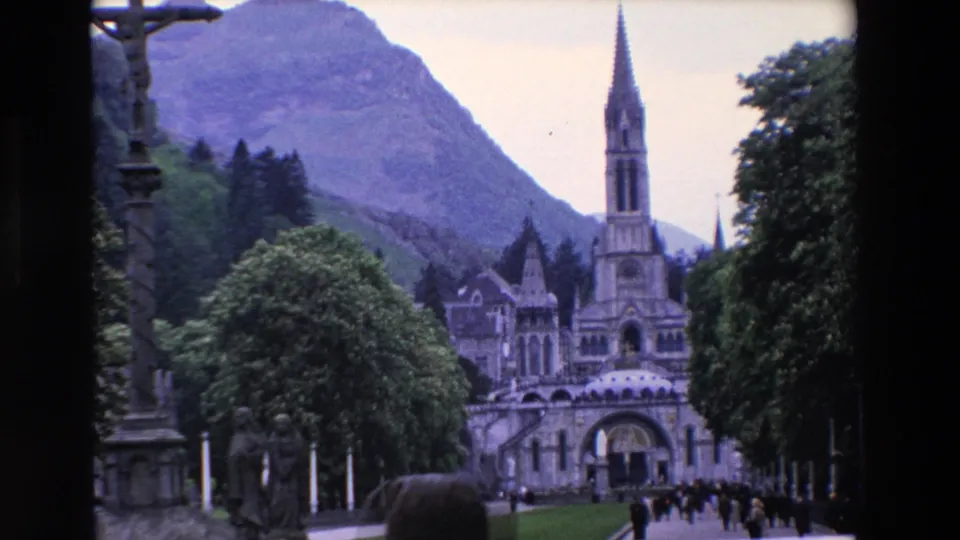 1969 8mm travel footage frame of a French basilica approach, with a tall central spire, stone facade, trees, mountain backdrop, and visitors walking near the entrance.