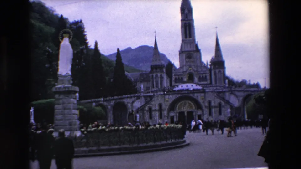 1969 French landmark footage showing a white religious statue on a pedestal, hedged walkway, groups of visitors, and the basilica facade with spires in the background.