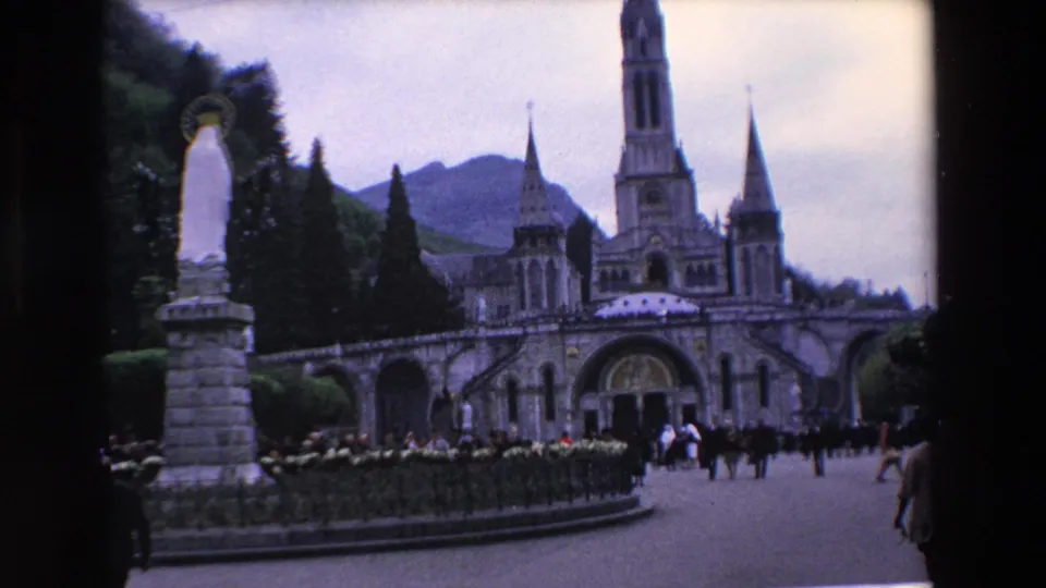 Visitor-shot 8mm frame from 1969 showing a statue and formal hedges leading toward a French basilica, with scattered pedestrians and mountains behind the roofline.