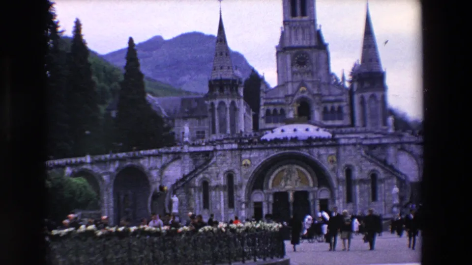Closer 1969 8mm frame of a French basilica facade, showing the arched entrance, tall spire, side towers, hedged foreground, visitors, and mountain backdrop.