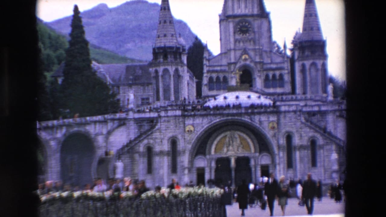 1969 8mm frame of a large stone religious building in France, cataloged as Paris but visually showing a mountainous backdrop, with visitors gathered across the courtyard and entrance steps.