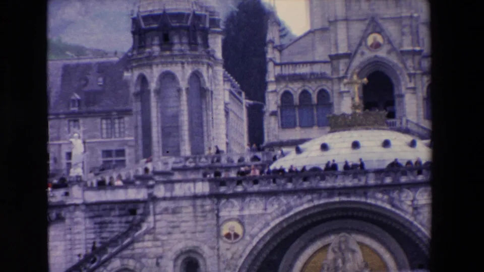 1969 8mm frame emphasizing the upper architecture of a large stone religious complex, including tower structures, terrace railings, a dome edge, and visitors standing along the balcony.