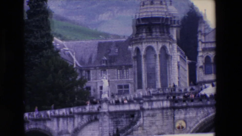 1969 8mm frame showing a partial side view of the church complex with a tower, crowded upper terrace, dark trees, adjacent stone building, and green hillside in the background.