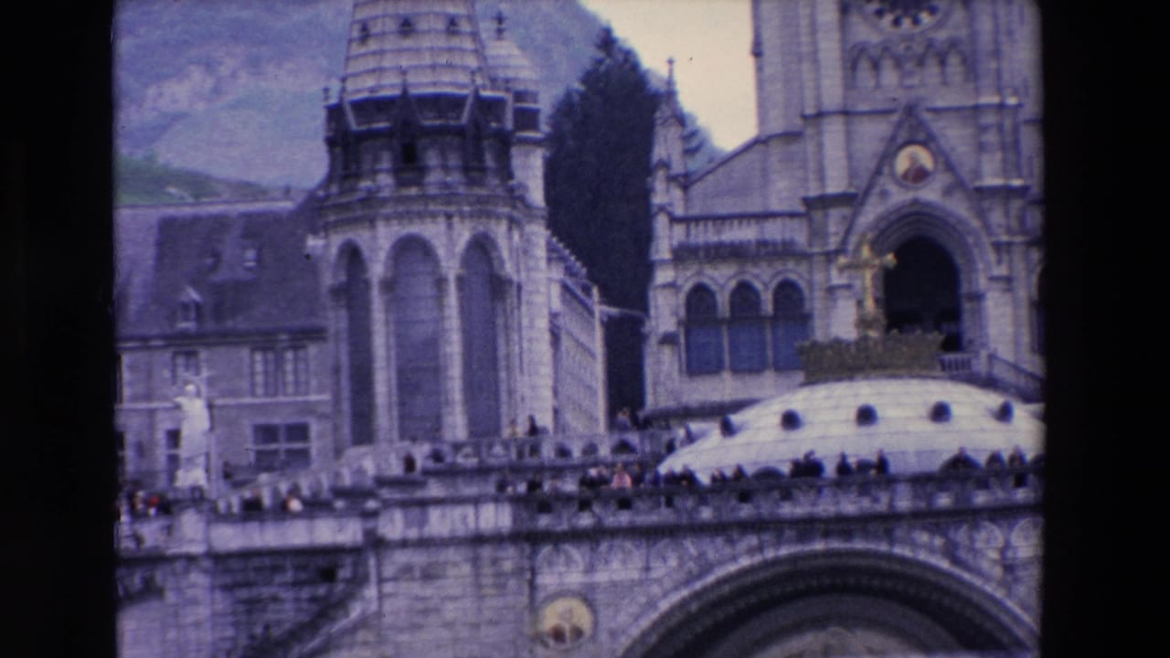 1969 8mm frame of a large French basilica facade, with stone towers, arched openings, terrace visitors, trees, and mountains in the background.