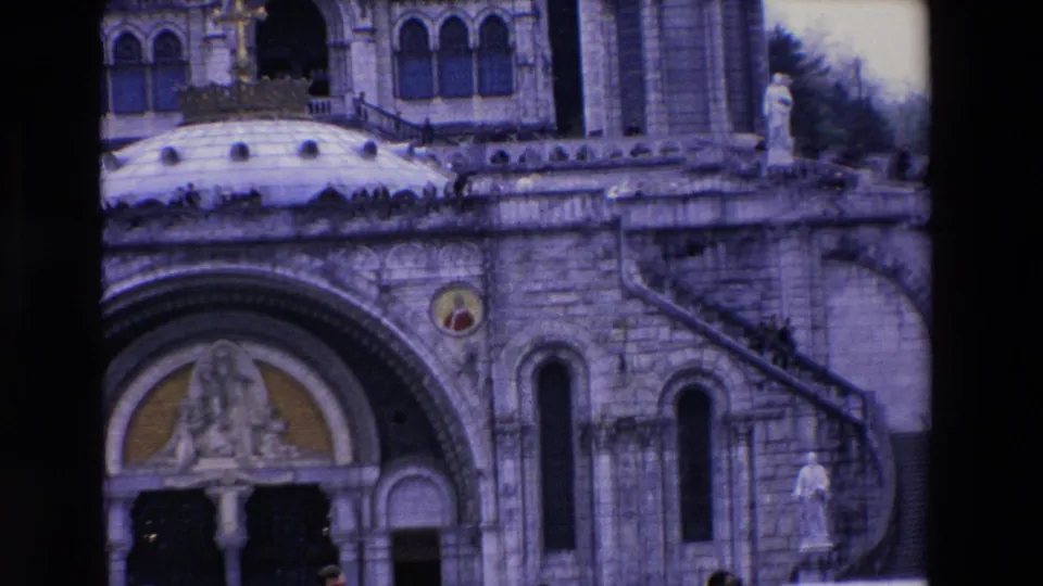 1969 8mm frame of the lower basilica exterior, with an arched doorway, religious sculpture, stone steps, terrace walls, and a statue on the side.