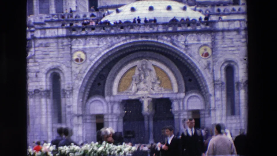 1969 French home-movie frame showing the basilica entrance below a religious sculpture, with flowers in the foreground and tourists gathered near the doorway.