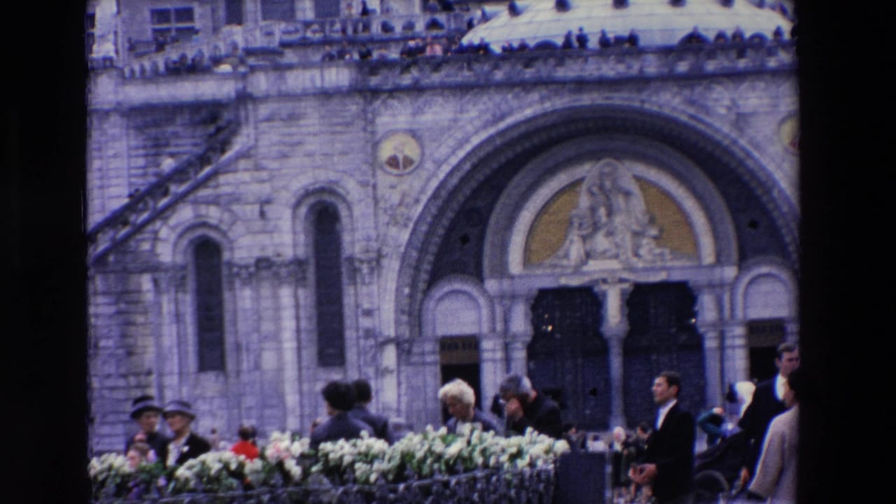 1969 Paris home-movie frame showing visitors outside a stone church or shrine entrance, with floral arrangements and people in period dress near the doorway.