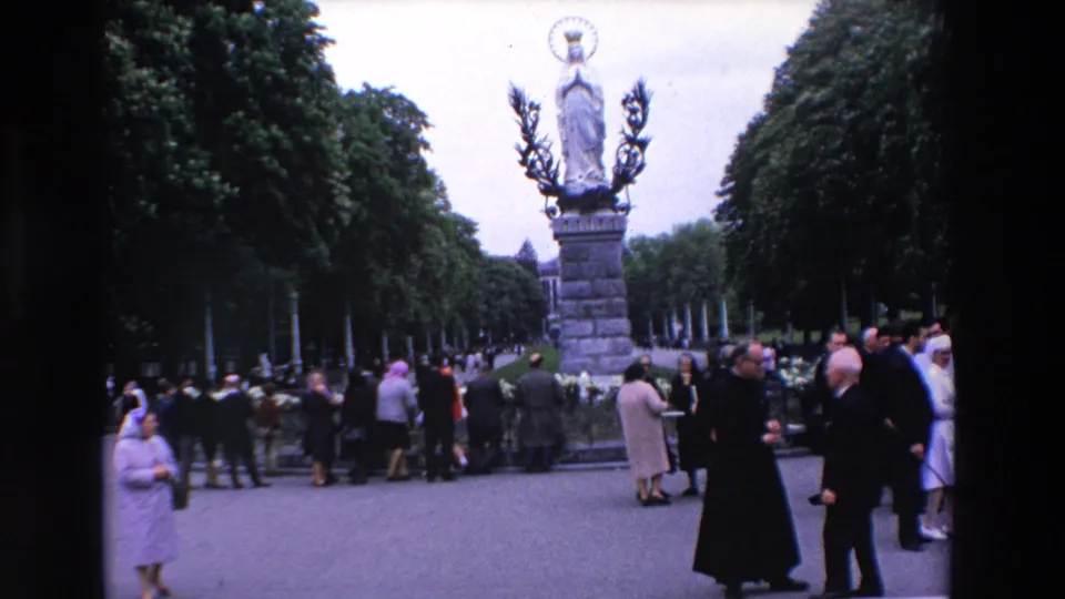 1969 Paris home-movie frame showing a Marian statue on a tall pedestal with priests and visitors gathered along a tree-lined walkway.