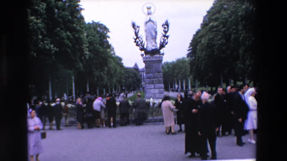 1969 Paris home-movie frame showing clergy and visitors gathered near a Marian statue, with rows of trees framing the plaza in the background.