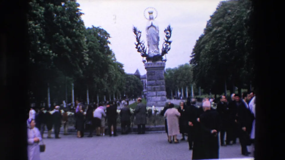 1969 Paris home-movie frame showing a crowd around a Marian statue on a pedestal, with priests and visitors visible in a tree-lined public religious setting.