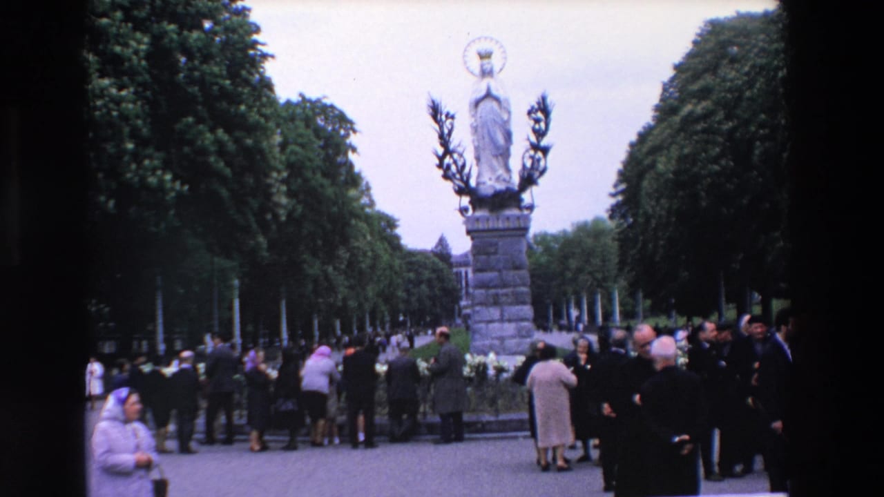 1969 Paris home-movie frame showing adults gathered near a crowned Virgin Mary statue on a stone pedestal in a tree-lined outdoor setting.