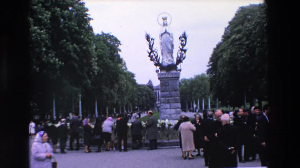 1969 Paris home-movie frame showing adults gathered near a crowned Virgin Mary statue on a stone pedestal in a tree-lined outdoor setting.