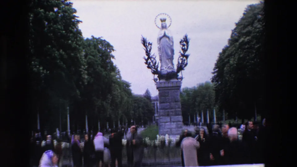 1969 Paris frame with the Virgin Mary monument centered above a small crowd gathered along a shaded tree-lined path.