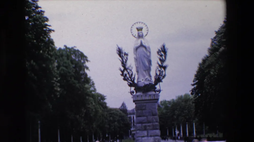 Closer 1969 Paris home-movie view of the crowned Virgin Mary statue and pedestal, with trees and a partially visible crowd below.