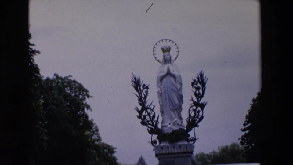 Tight 1969 Paris frame of the crowned Virgin Mary statue on a stone pedestal, framed by dark metal wreath forms, sky, and trees.