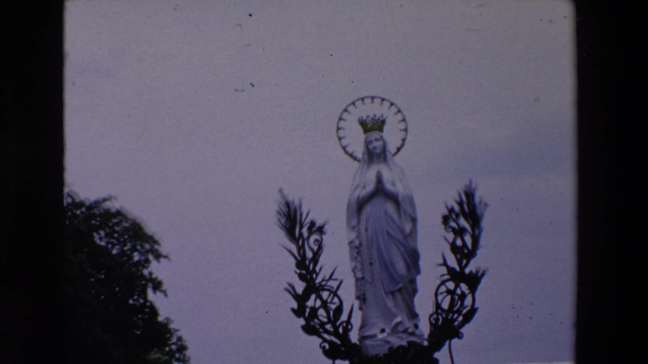 1969 Paris 8mm frame showing a white Virgin Mary statue with clasped hands, crown, and circular halo against a pale sky, partly framed by dark foliage and metalwork.