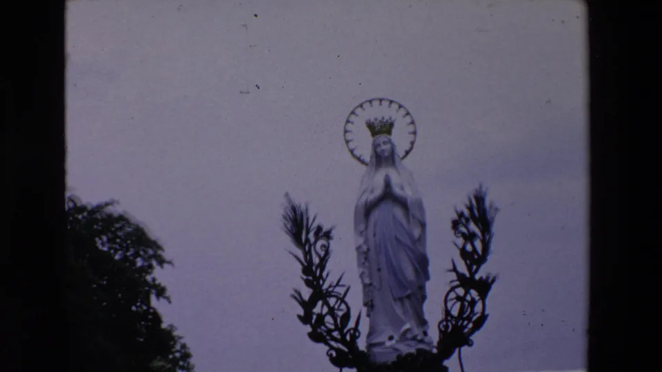 1969 Paris 8mm frame showing a white Virgin Mary statue with clasped hands, crown, and circular halo against a pale sky, partly framed by dark foliage and metalwork.