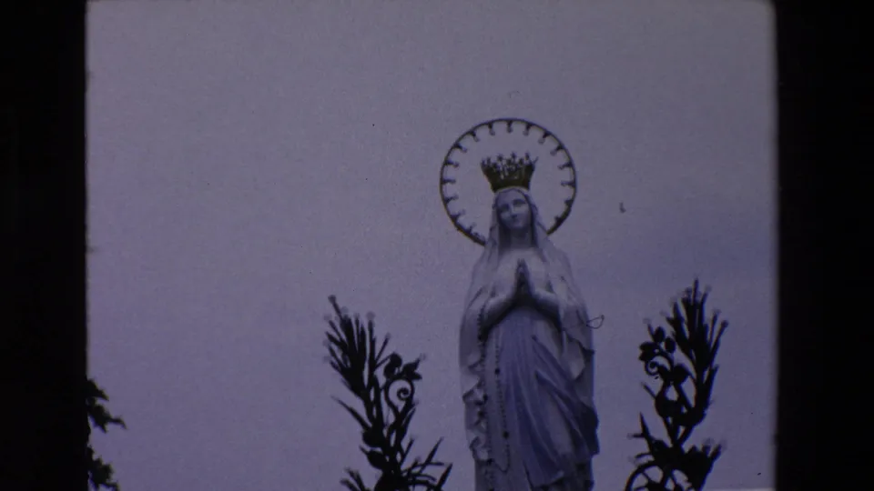 1969 Paris home-movie frame with the Virgin Mary statue more centered, showing the crown, halo, praying hands, and dark ornamental shapes at the sides.