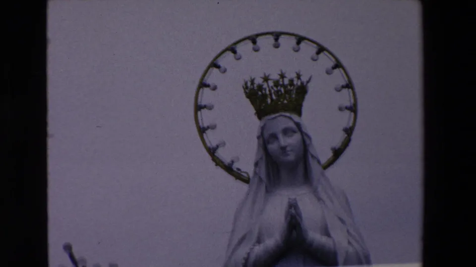 Tight 1969 Paris home-movie frame showing the Virgin Mary statue's face, crown, circular halo, and clasped hands against a plain pale sky.