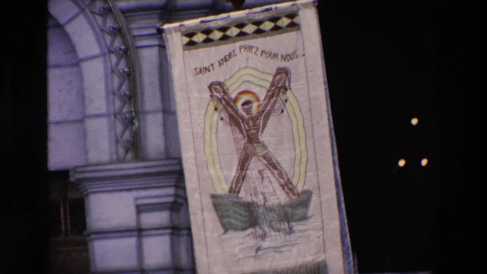 Close 8mm view of a religious banner reading Saint André Priez Pour Nous, suspended beside the ornate church doorway during the Paris crowd scene.