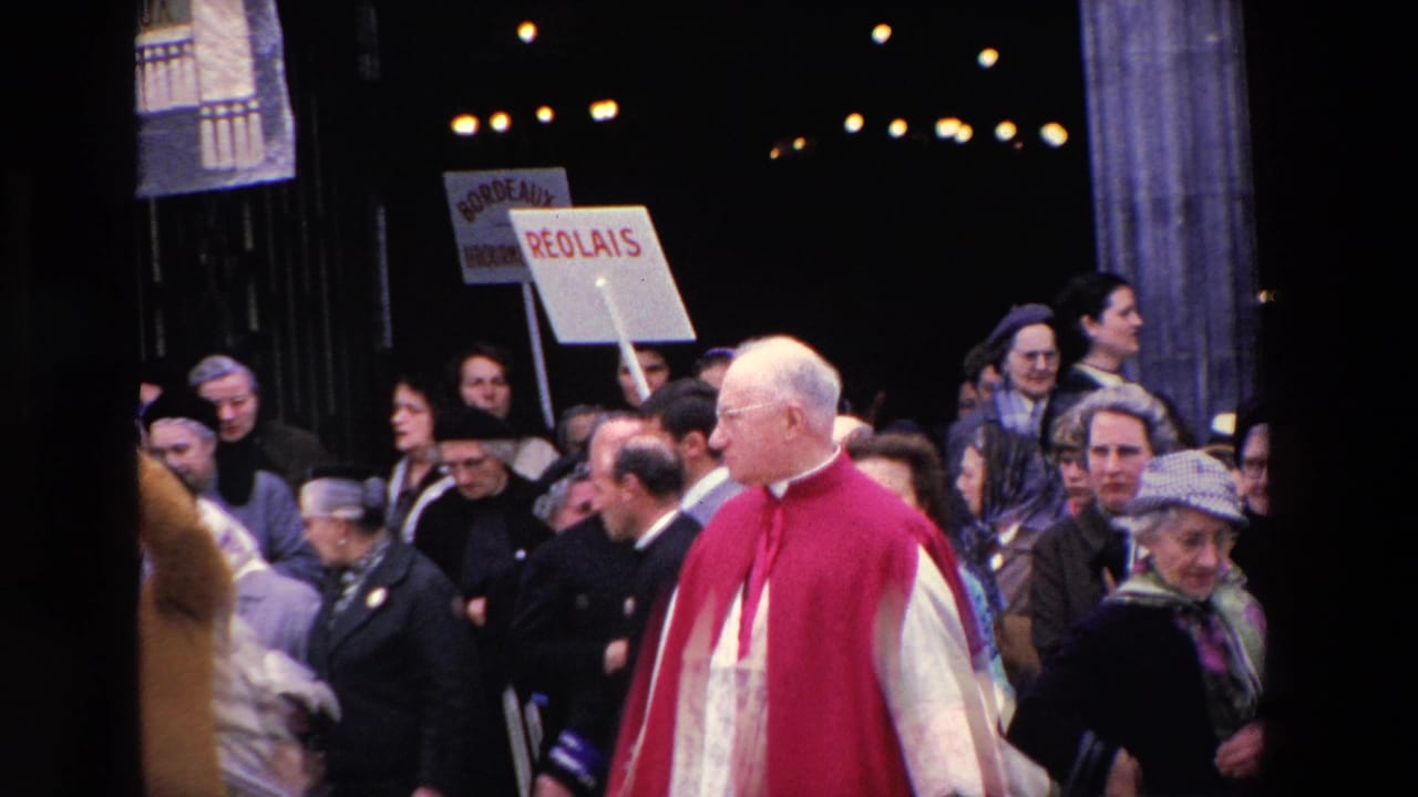 In Paris in 1969 according to clip metadata, a red-caped clergyman moves through a dense crowd while a placard with French place names is raised behind him.