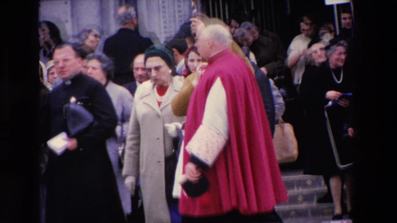 A red-caped clergyman appears near a stone entrance in the 1969 Paris street scene, surrounded by onlookers and another dark-robed religious figure.